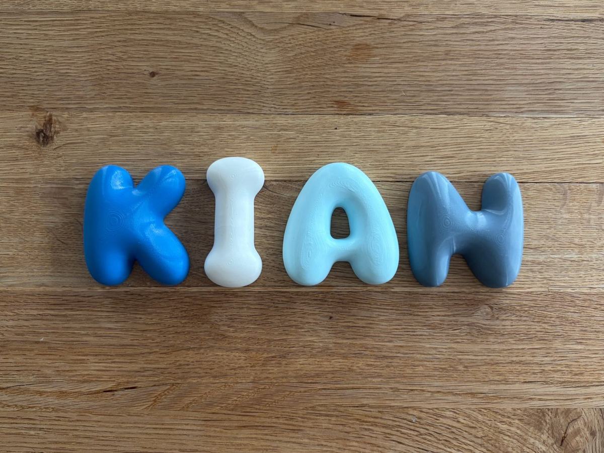 Die Buchstaben K, I, A und N in Blau, Weiss, Hellblau und Graublau auf einem Holztisch